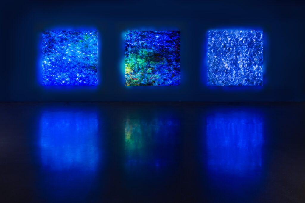 Three blue square paintings lit from behind in dark gallery.